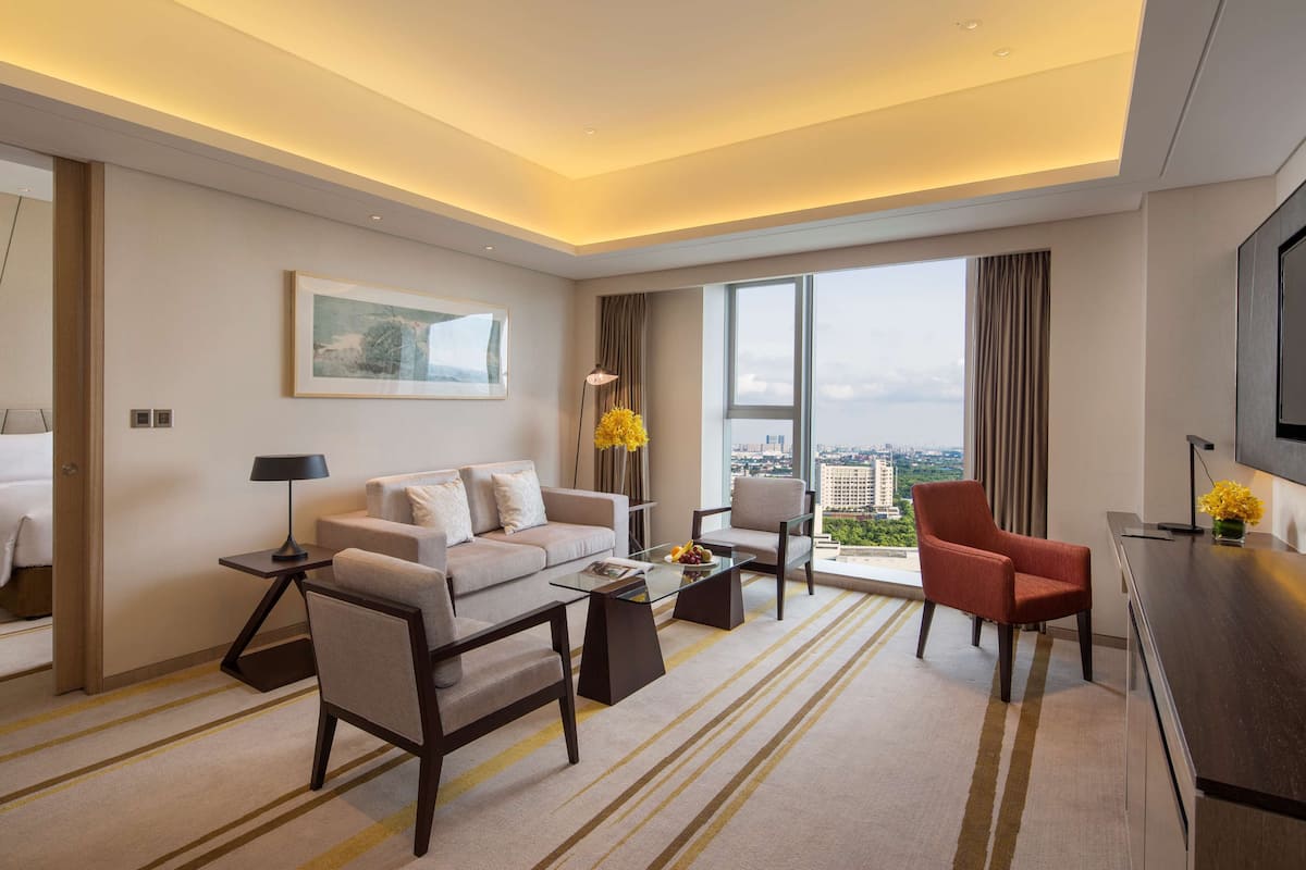 A modern hotel room with a sofa, armchair, coffee table, and a desk with a TV.
