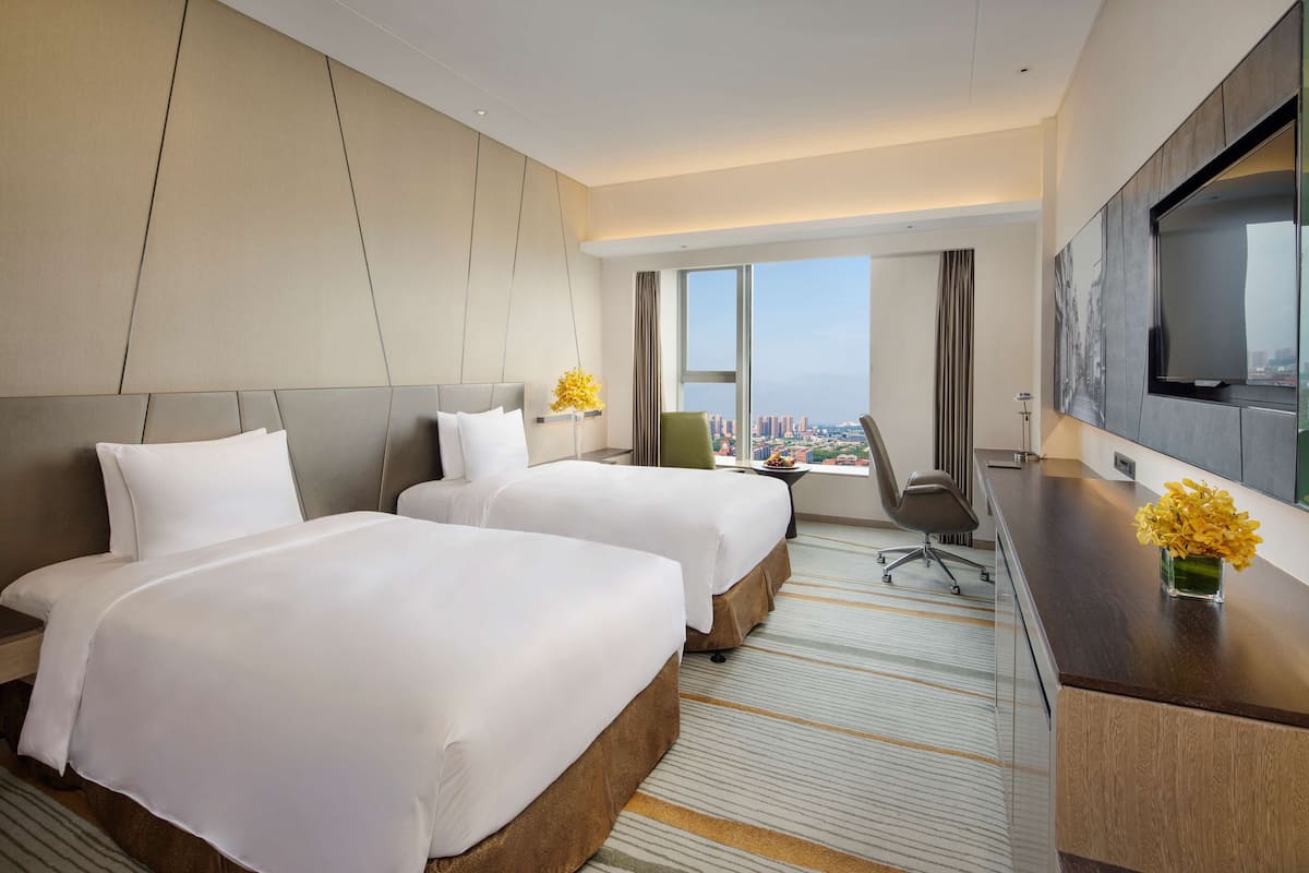 A hotel room with two beds, a desk, a chair, a TV, and a large window offering a city view.