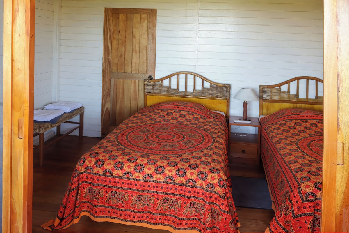 Een kamer met twee houten bedden met beddengoed met patroon, een houten bankje, een klein tafeltje met een lamp en een houten deur.