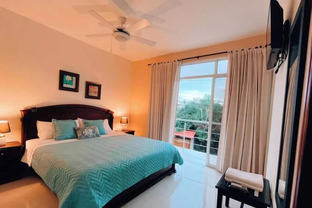 Quarto de hotel com cama, duas luminárias laterais, ventoinha no teto, janela com cortinas e uma pequena mesa com um controle remoto.