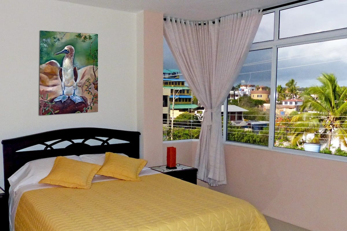 Quarto com cama, mesa de cabeceira, um quadro e janela com vista para edifícios e palmeiras.