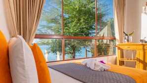 Habitación de hotel con un ventanal grande, una cama con ropa de cama blanca y naranja, y una mesita de noche de madera con una botella de champán.