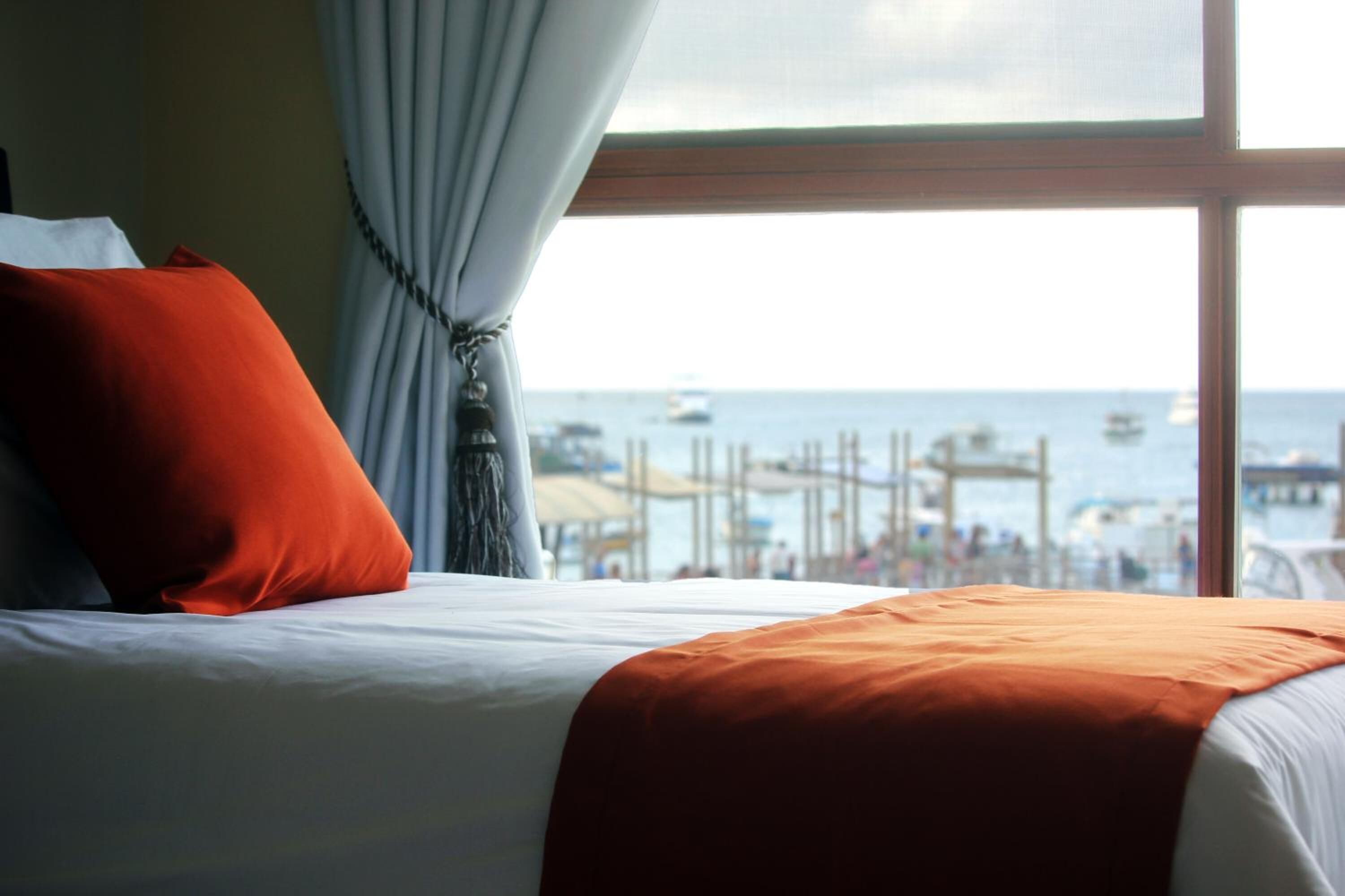 A bed with white bedding and an orange throw blanket, a red pillow, and a view of the sea and boats through a window.