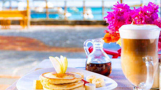 Desayuno con tortitas, plátanos, almíbar y una bebida de café sobre una mesa con vista al mar.