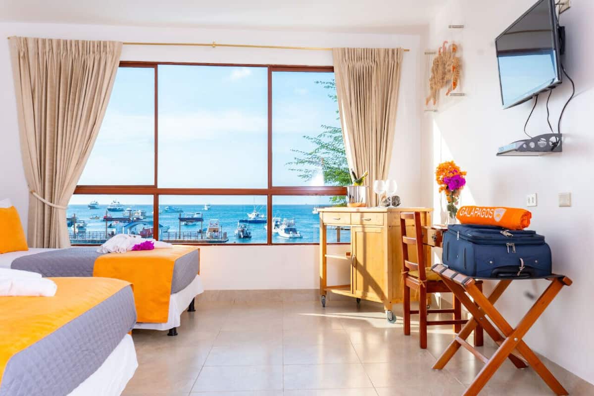 A hotel room with two beds, a desk, a chair, a TV, and a view of the sea.