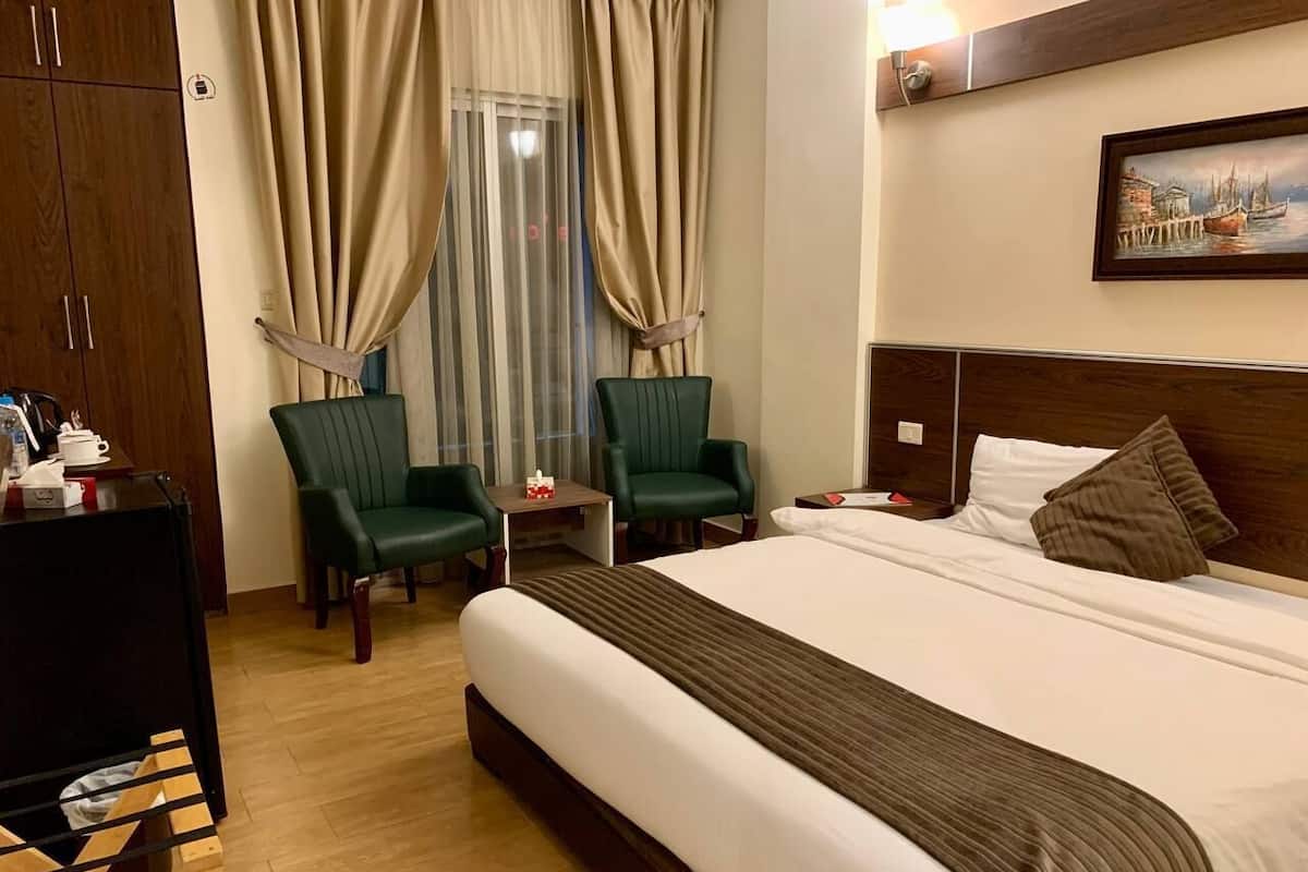 A hotel room with a large bed, two green armchairs, a small table, and a wooden wardrobe.
