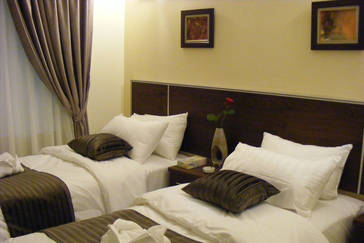 A hotel room with two beds, each with white bedding and striped pillows. There is a vase with a single rose on the nightstand between the beds.