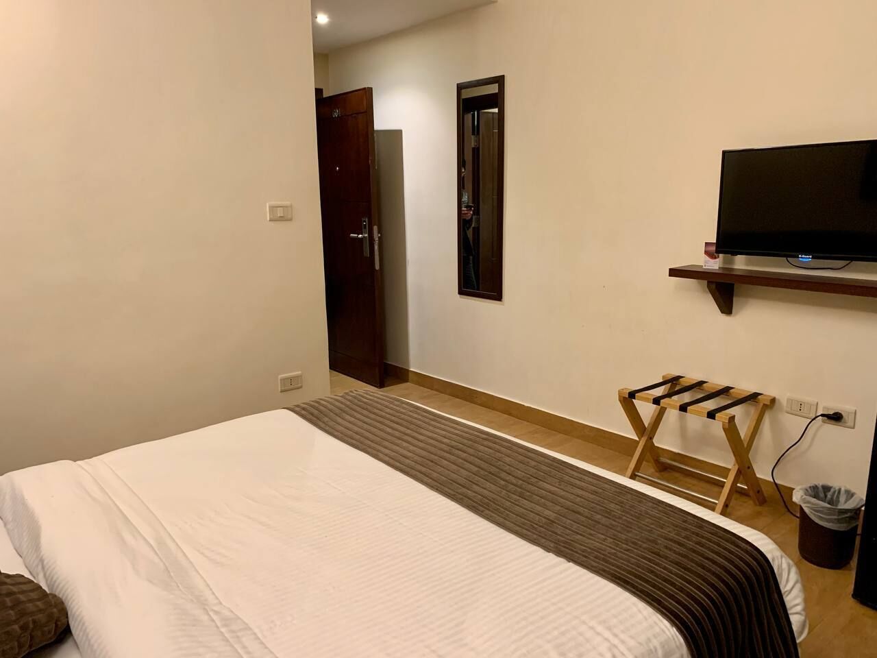 A hotel room with a bed, a TV on a stand, a wooden stool, and a trash bin.