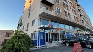 Un hotel de varios pisos con diseño moderno, que cuenta con balcones en cada planta, una entrada a nivel de calle y un pequeño quiosco azul.