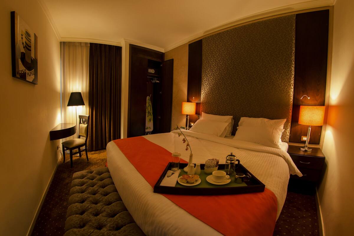 A hotel room with a large bed, a bedside table with a lamp, a chair, and a tray with breakfast items.