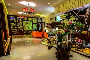 A modern lobby with a glass table, orange seating, decorative lighting, and a reception desk. - Meneur Hotel (Amman)