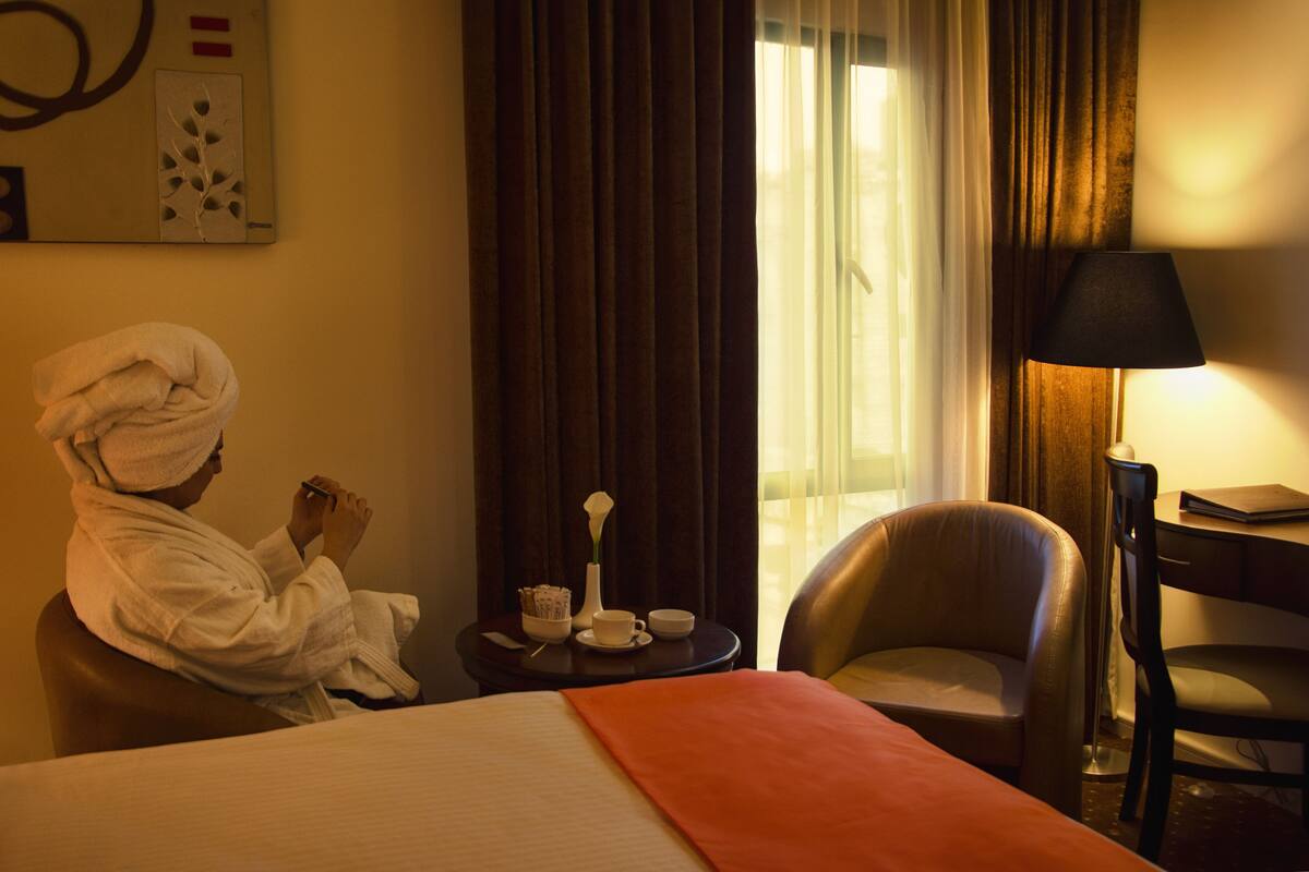 A hotel room with a bed, a chair, a table with a lamp, and a window with curtains.