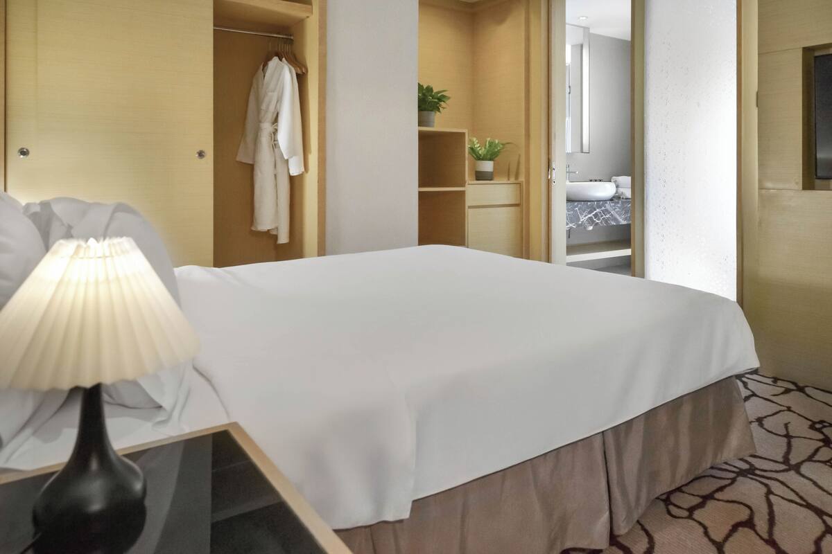 A hotel room with a bed, bedside lamp, wardrobe, and a bathroom visible through an open door.