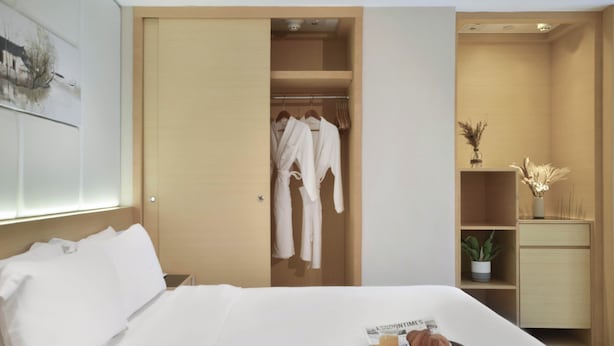 A hotel room with a neatly made bed, a tray with pastries, a wardrobe with robes, and a small plant on a shelf.