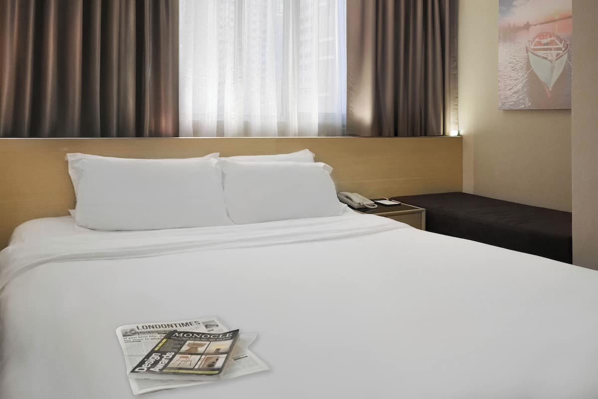 A hotel room with a neatly made bed, a bedside table with a lamp, and a window with curtains.