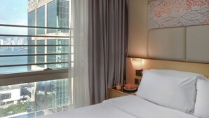A hotel room with a large bed, a nightstand with a lamp, a view of the city through a large window, and a cityscape visible outside.