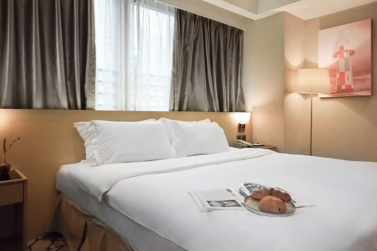 A neatly made bed with white linens, a bedside lamp, and a newspaper with pastries on the bed.