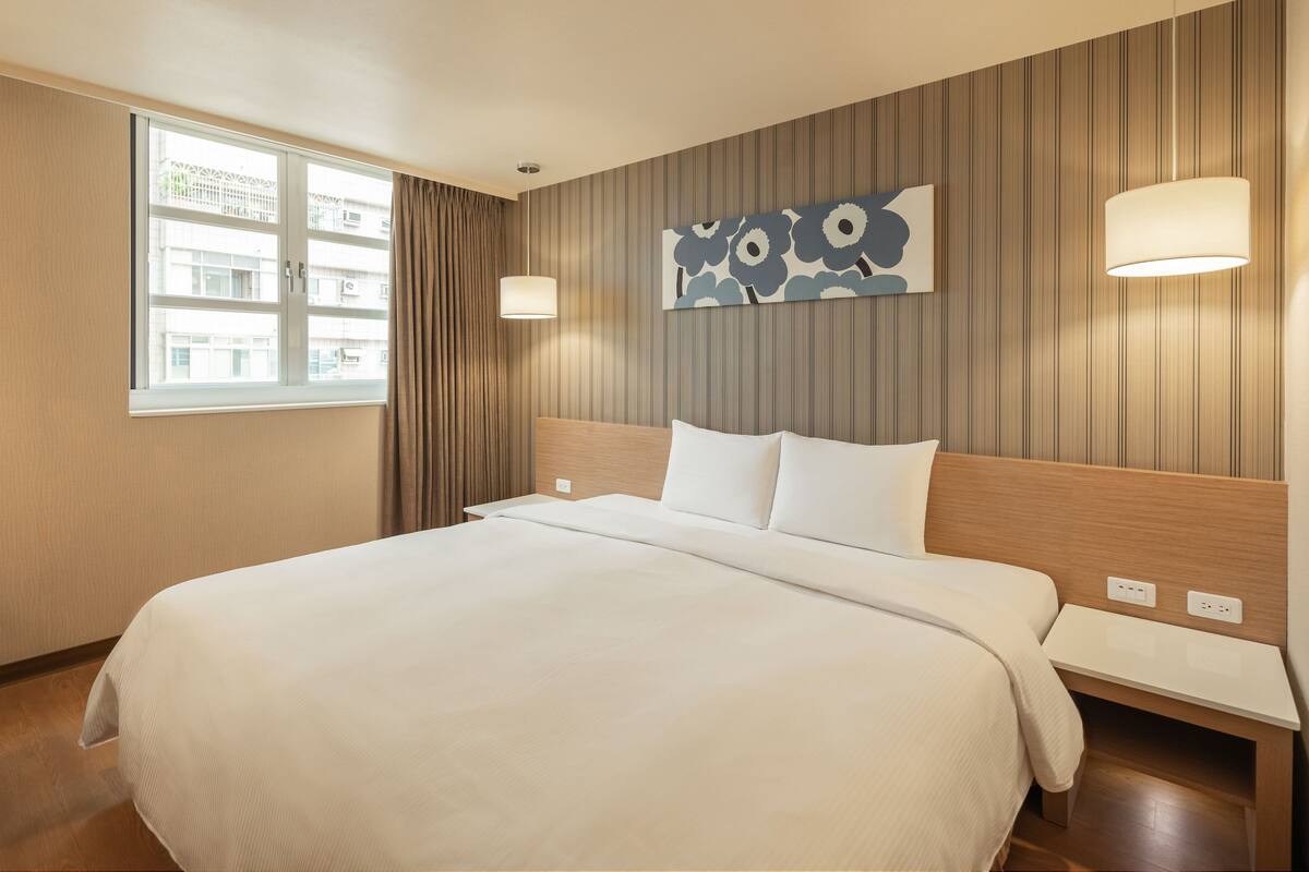 Une chambre d’hôtel avec un grand lit, une fenêtre avec des rideaux, une œuvre d’art accrochée au mur et une table de chevet avec une lampe.
