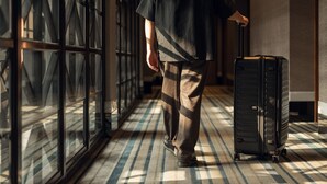 Une personne marchant dans un couloir, une valise à la main, avec un sol à motifs et de grandes fenêtres.