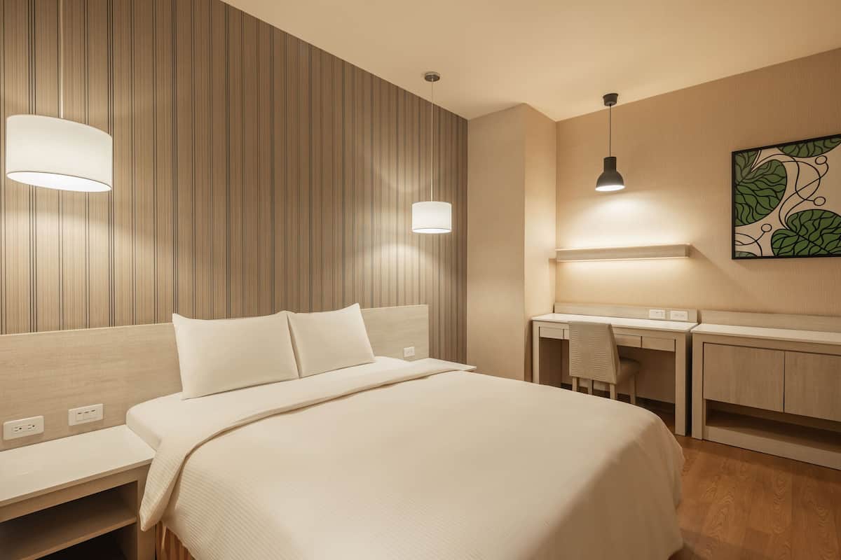 Une chambre d’hôtel avec un lit, des tables de chevet, un bureau et une chaise. Il y a deux lampes fixées au mur et un tableau accroché au mur.