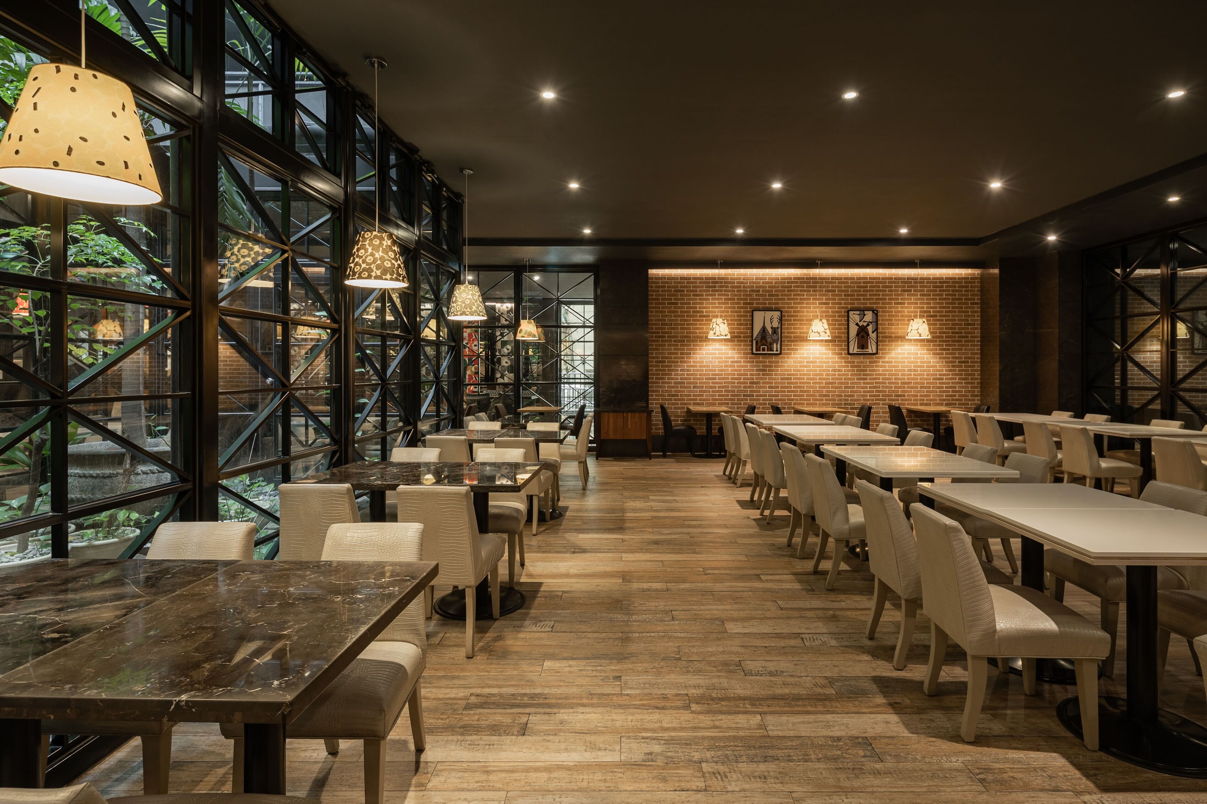 A restaurant with high ceilings, large windows, and a brick wall. Tables and chairs are arranged for dining.