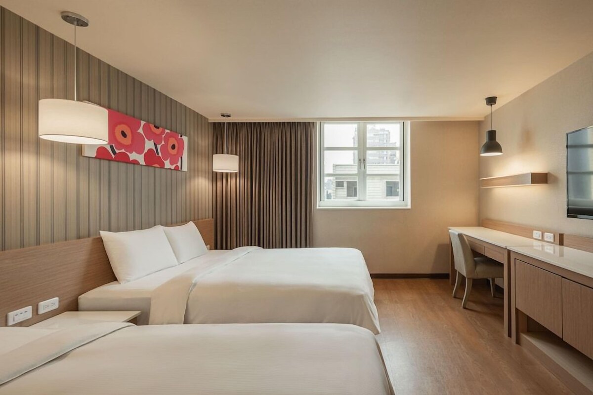 Habitación de hotel con dos camas, un escritorio, una silla, un televisor, una ventana y un adorno en la pared.