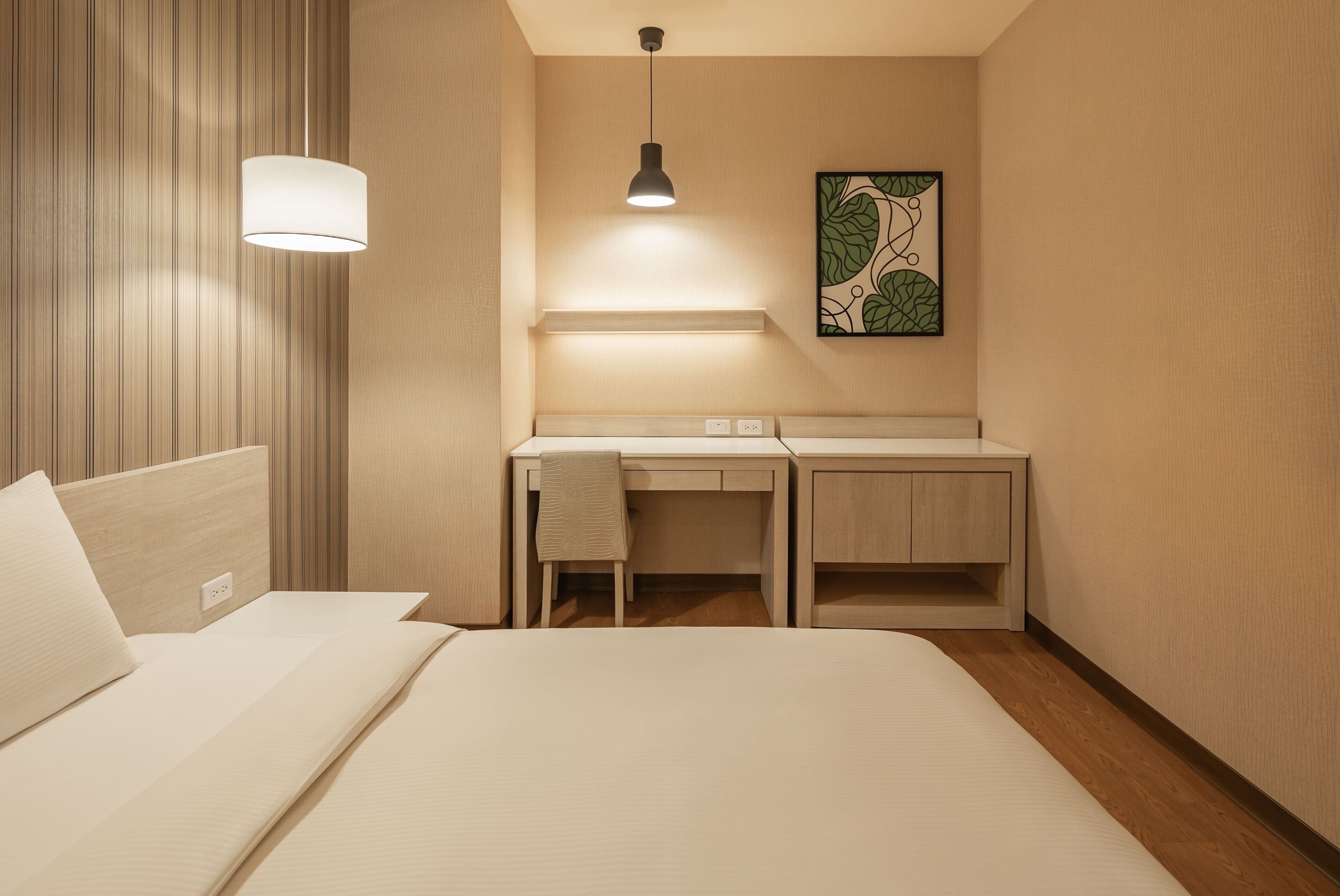 A hotel room with a bed, a desk, a chair, a hanging light, and a wall-mounted artwork.