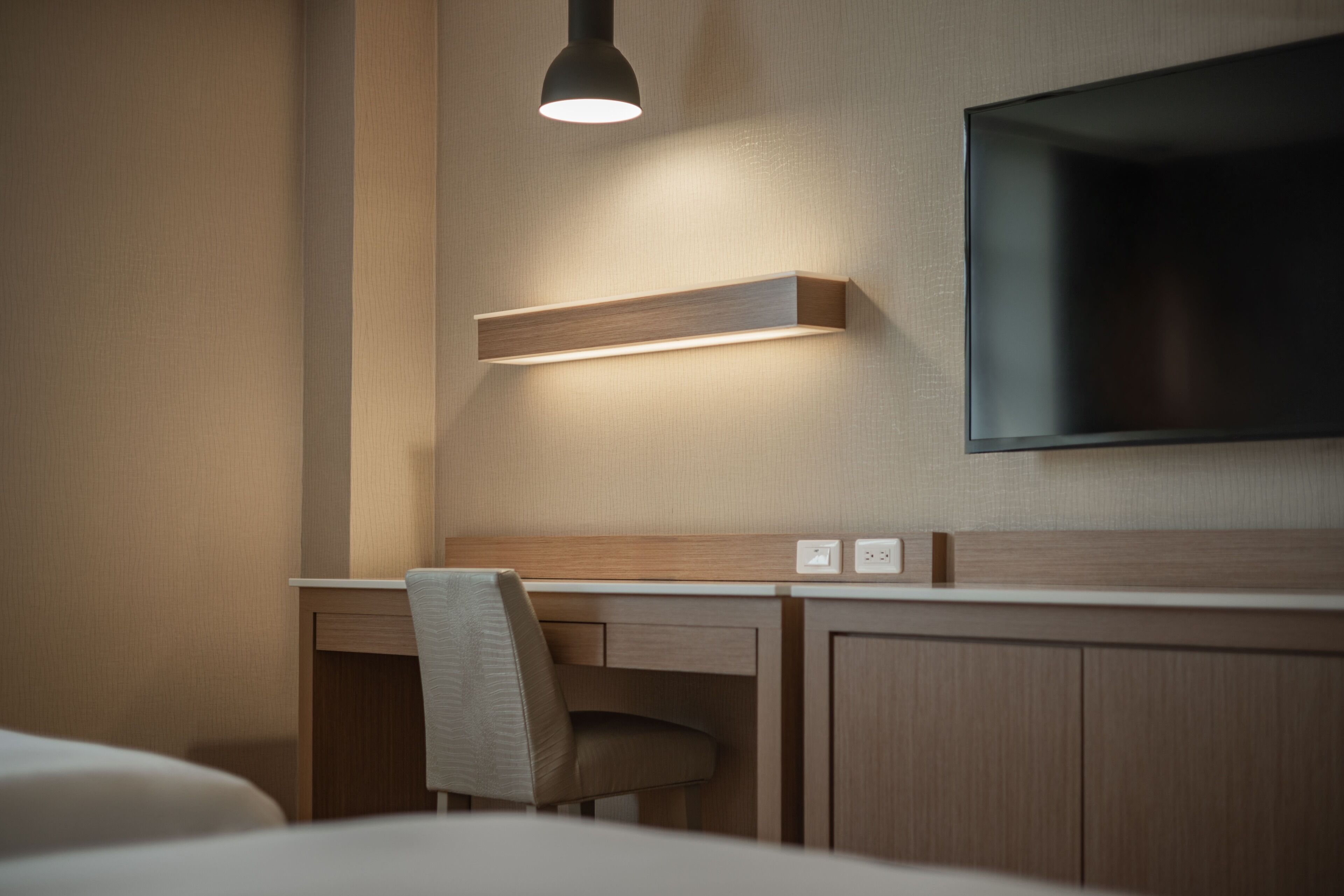 A hotel room with a bed, a desk, a chair, a TV mounted on the wall, and a light fixture above the desk.