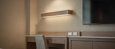 A hotel room with a bed, a desk, a chair, a TV mounted on the wall, and a light fixture above the desk.