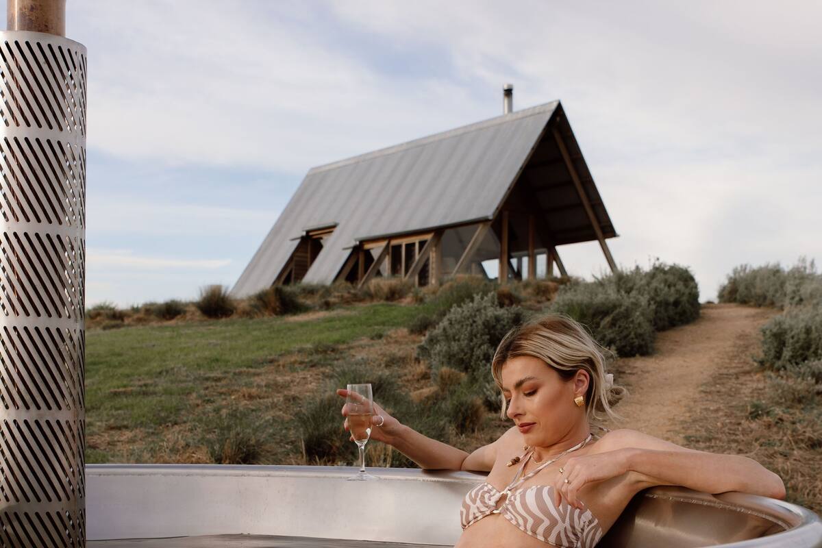 A person relaxing in a hot tub with a glass of wine, surrounded by a rustic wooden structure and a clear sky.