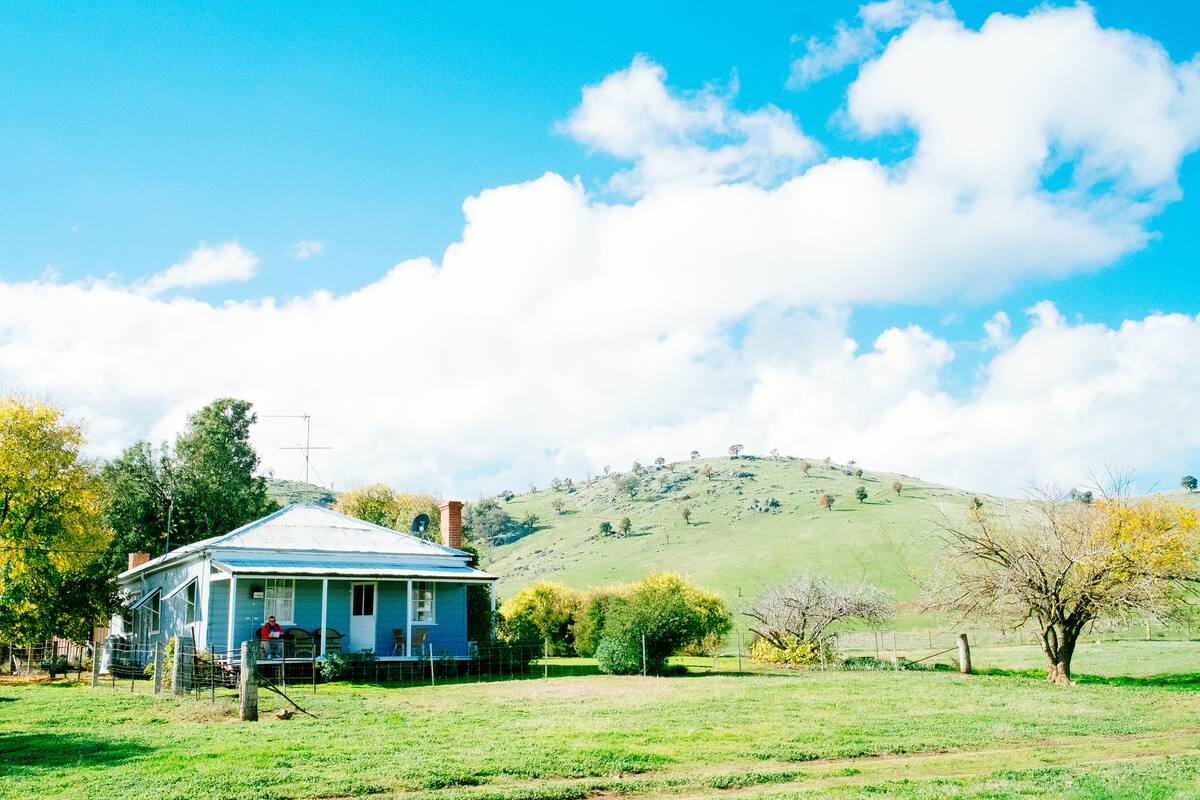 A house with a blue roof and a porch, surrounded by a green field and trees.