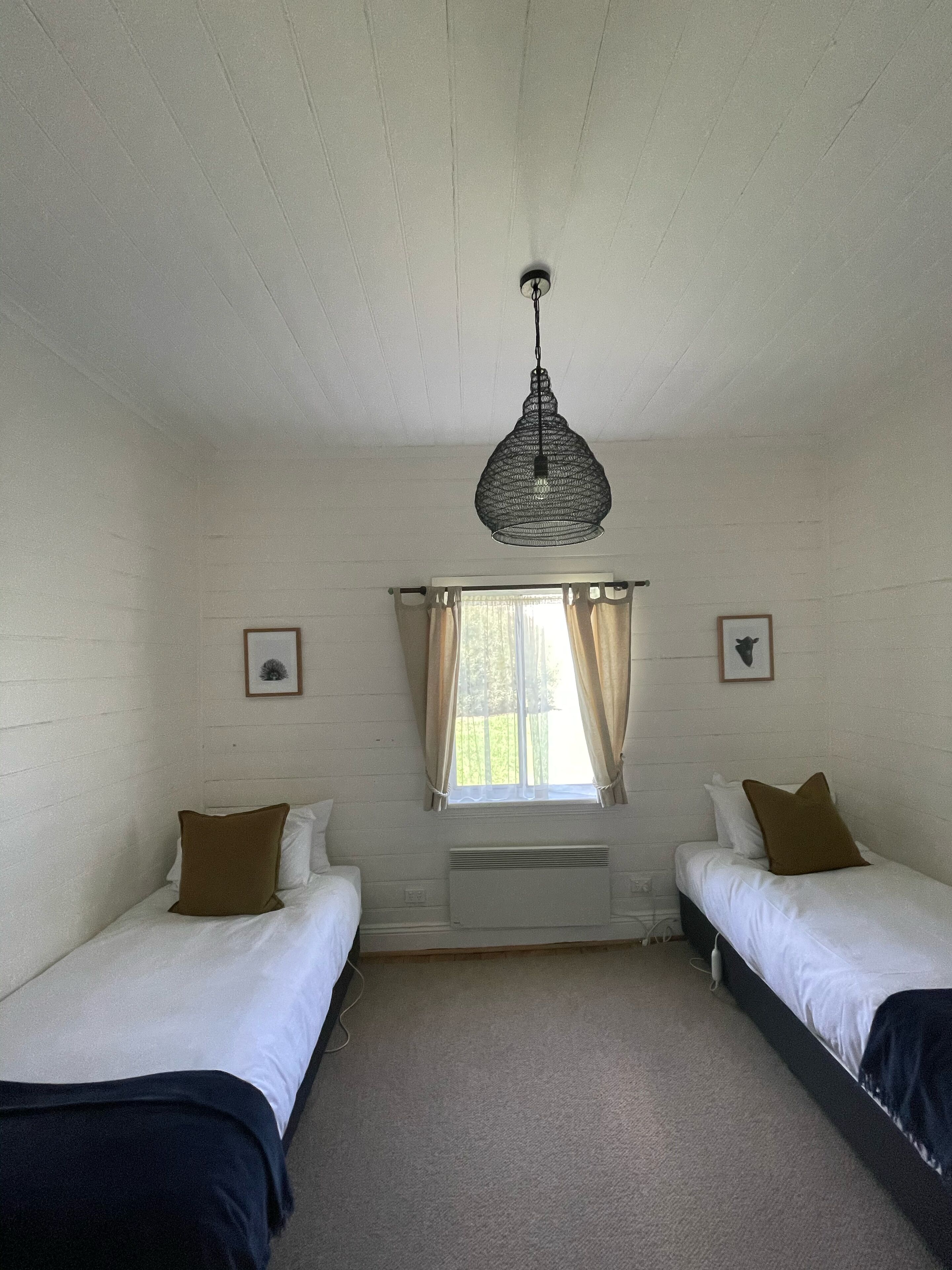 A room with two single beds, a hanging light fixture, a window with curtains, and a radiator.