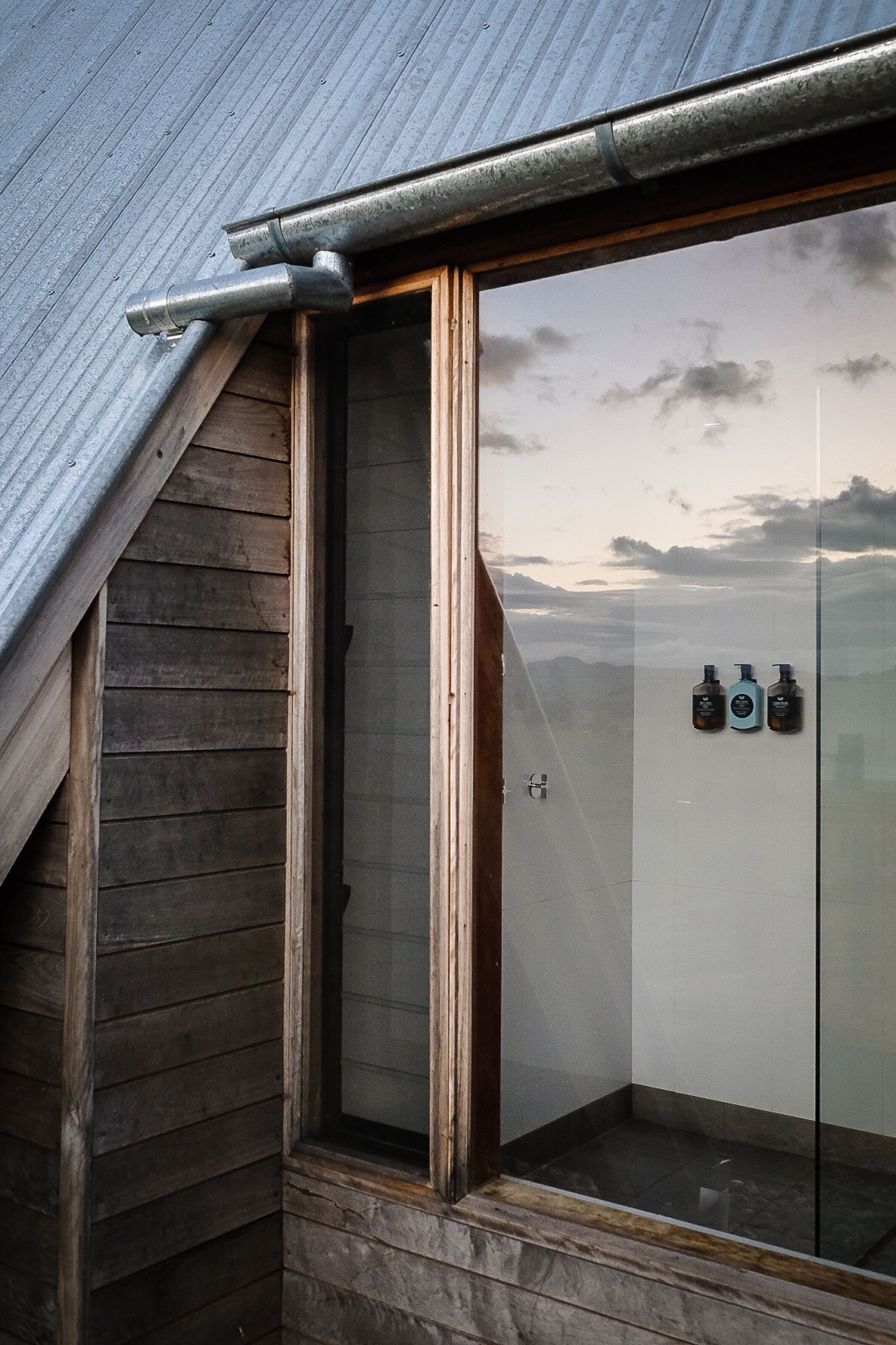 A corner of a building with a wooden exterior and a glass door leading to a room with a view of the sky.