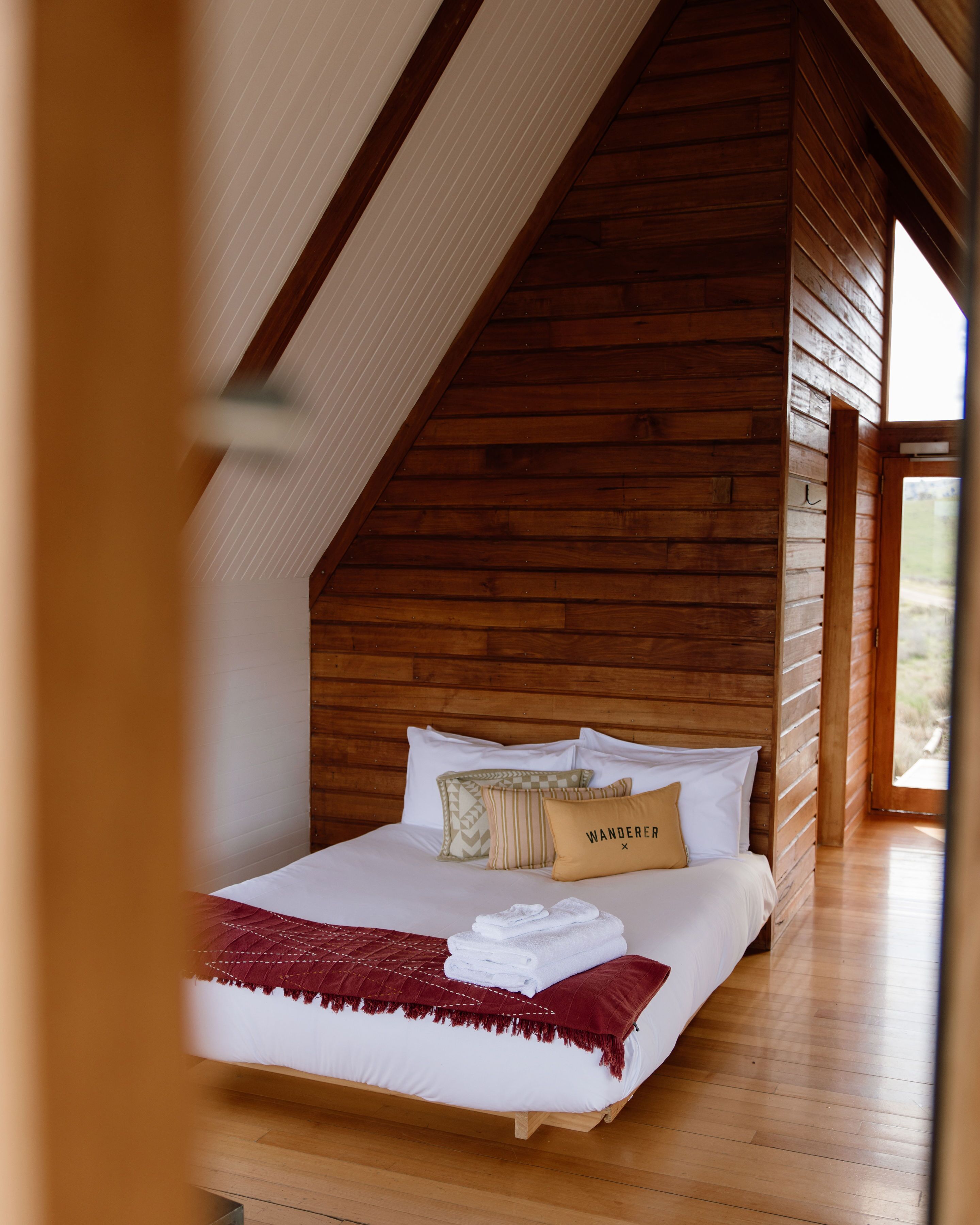 A wooden cabin-style bedroom with a sloped ceiling, a bed with white linens, a red throw blanket, and a pillow labeled "WANDERER."
