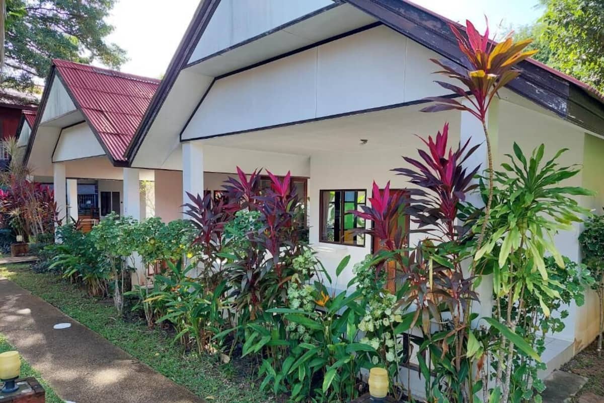 A modern building with a white facade and a red roof, surrounded by lush greenery and tropical plants.