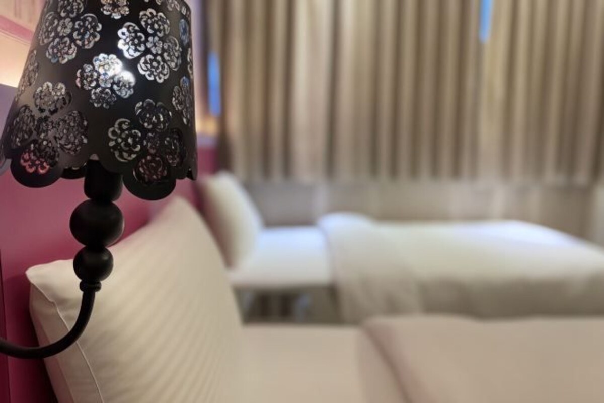 Primer plano de una lámpara con estampado floral, un almohadón blanco y una cama al fondo.