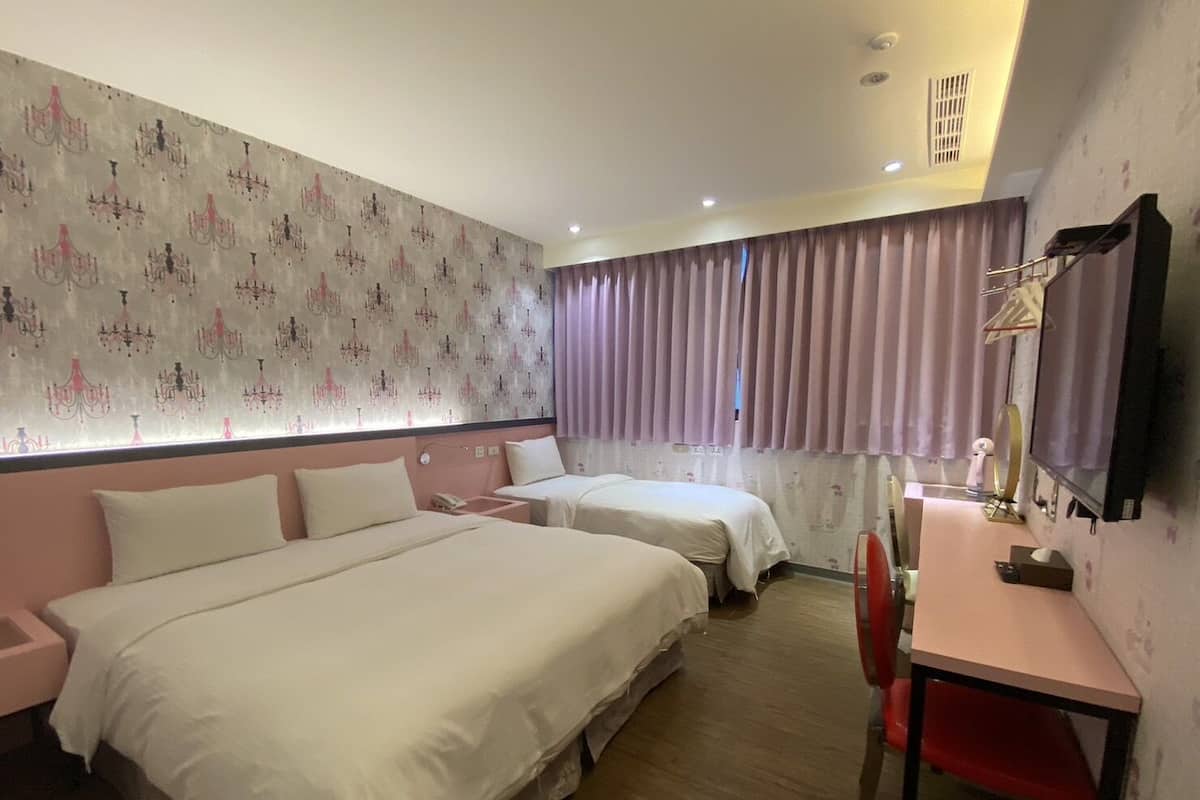 A hotel room with two beds, a desk, a chair, a TV, and a window with curtains.