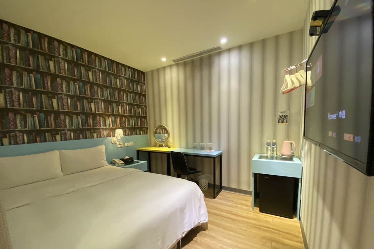 A hotel room with a large bed, a desk with a computer, a TV, and a bookshelf.