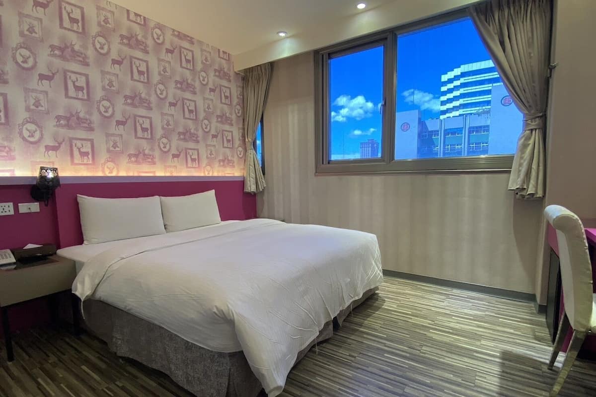 A hotel room with a large bed, a nightstand, a desk, and a window with a city view.