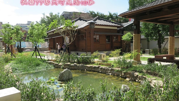 A traditional Japanese-style building with a wooden exterior and tiled roof, surrounded by a garden with a pond and various plants.