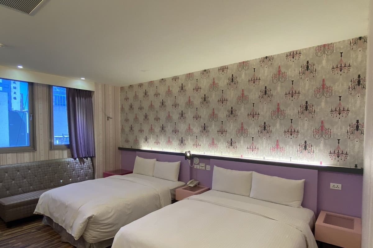 A hotel room with two beds, a large window with curtains, a patterned wallpaper, and a built-in bench.