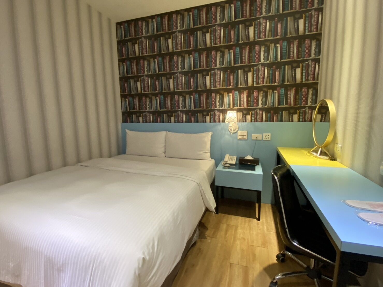Ein Hotelzimmer mit einer großen Bücherwand, einem Bett, einem Schreibtisch mit Stuhl sowie einem Nachttisch mit Lampe.