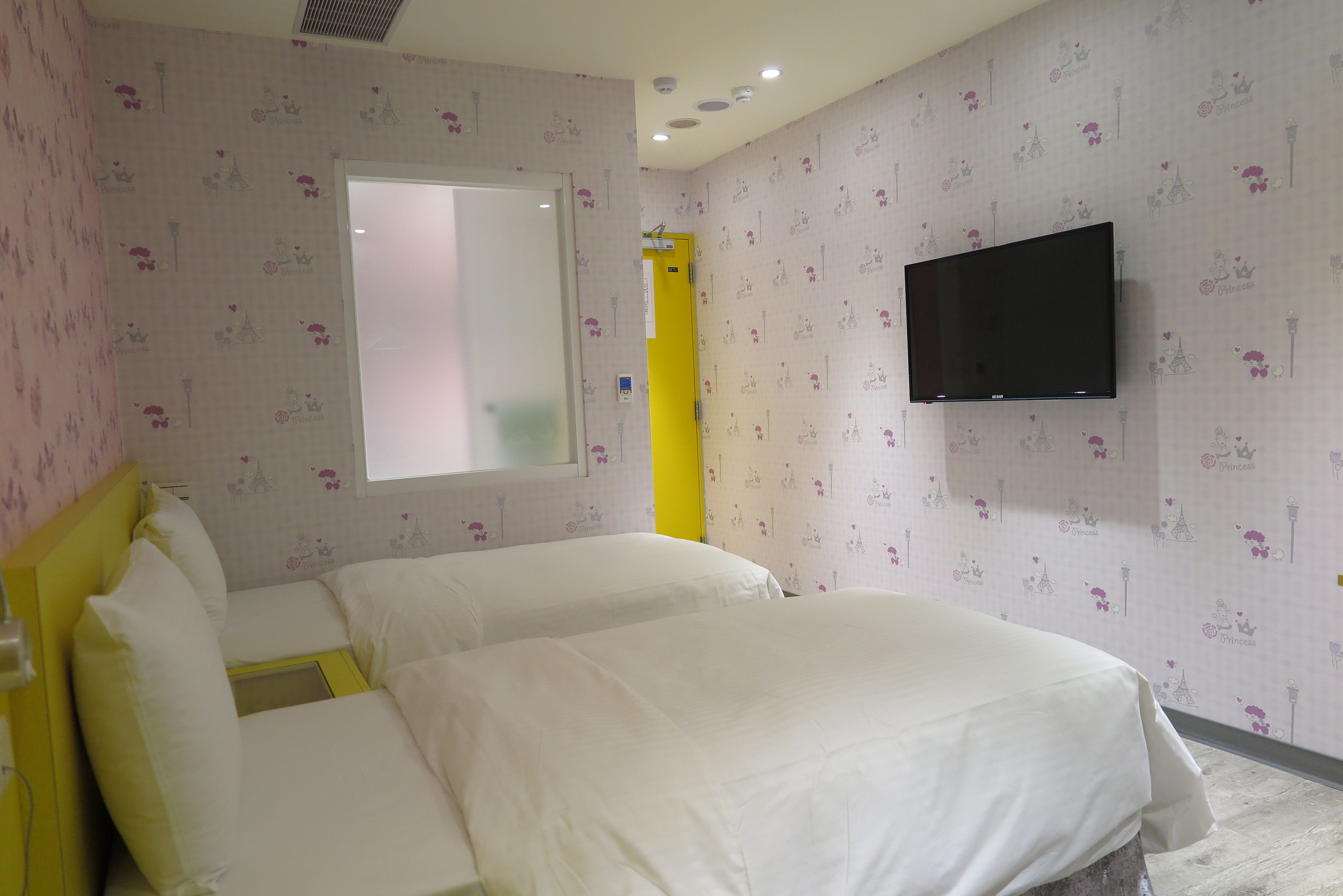 A hotel room with two beds, a flat-screen TV mounted on the wall, and a window with a view of the city.
