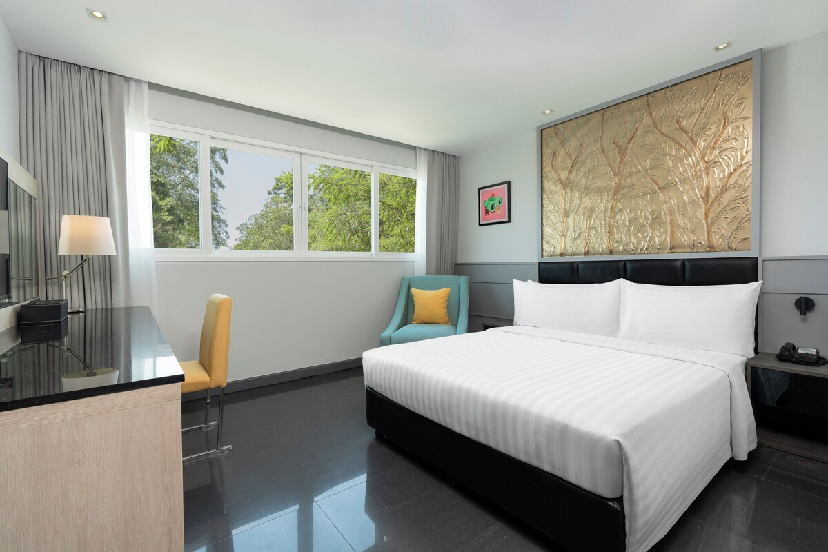 Una habitación de hotel moderna con una cama grande, un escritorio con lámpara, una silla, una mesita de noche y una ventana con vista a árboles.