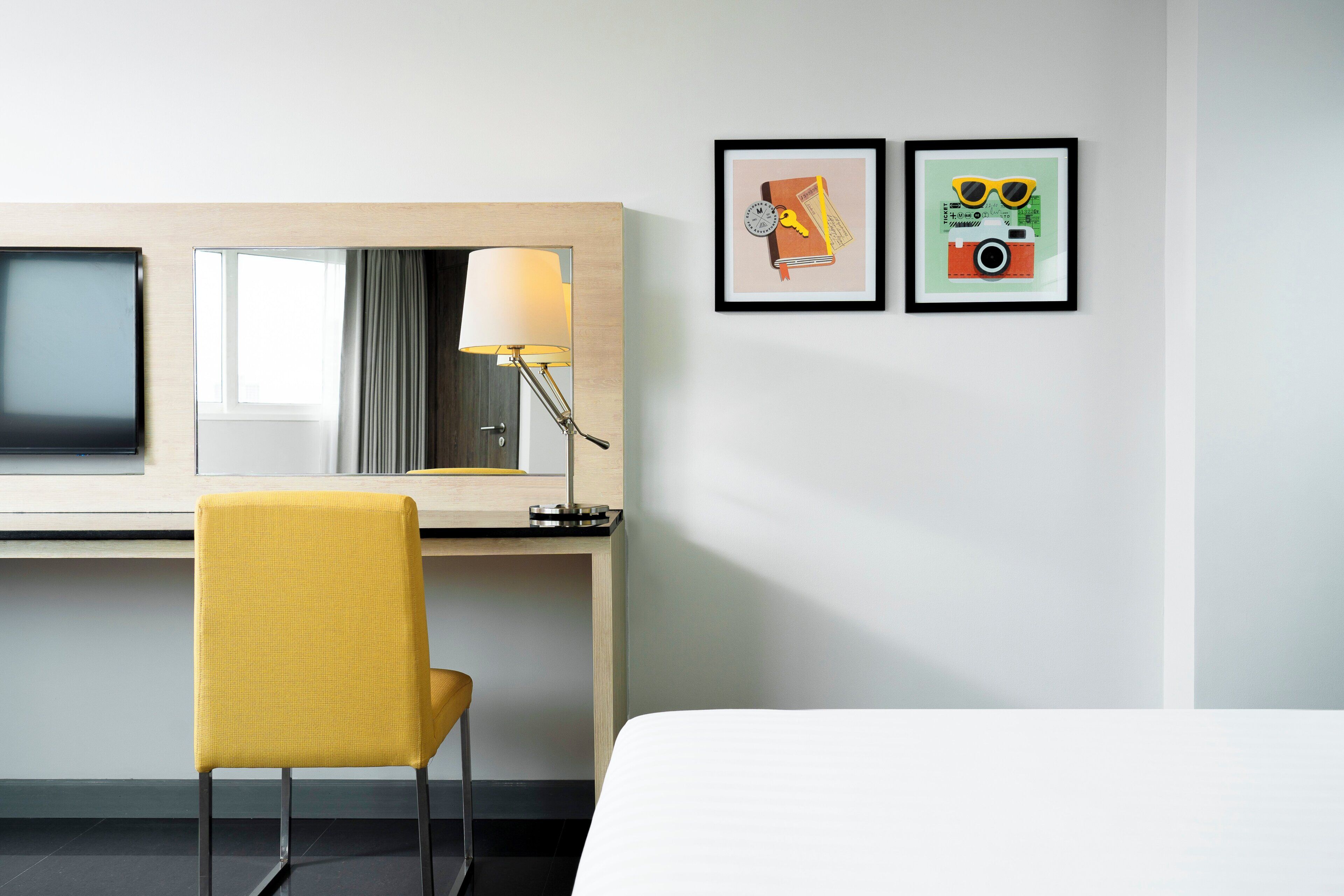 A hotel room with a bed, a desk with a lamp, a chair, and two framed pictures on the wall.