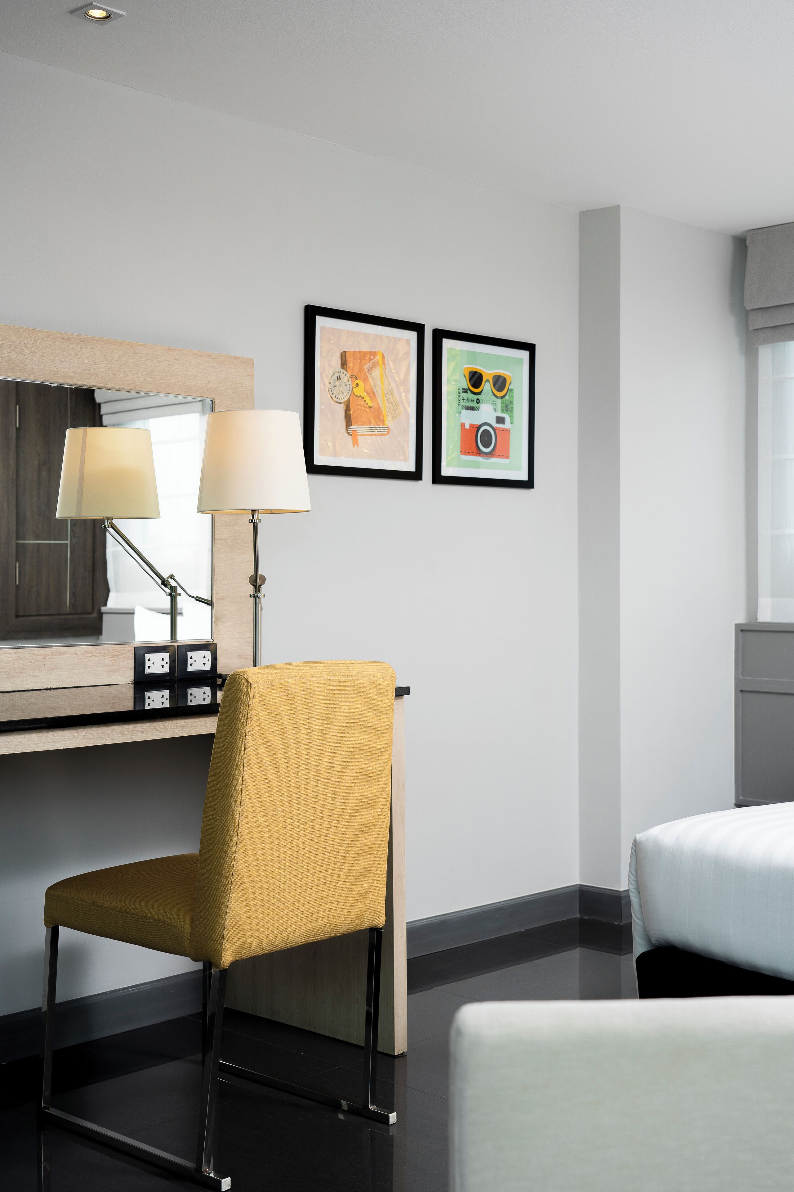 A hotel room with a desk, two lamps, a chair, a bed, and two framed pictures on the wall.
