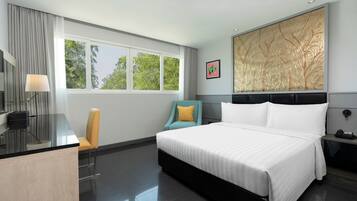A modern hotel room with a large bed, a desk with a lamp, a chair, and a view of trees through the window.