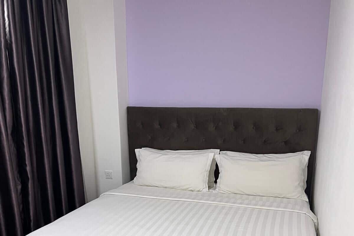 A single-bed room with a dark headboard, white bedding, and a purple accent wall.