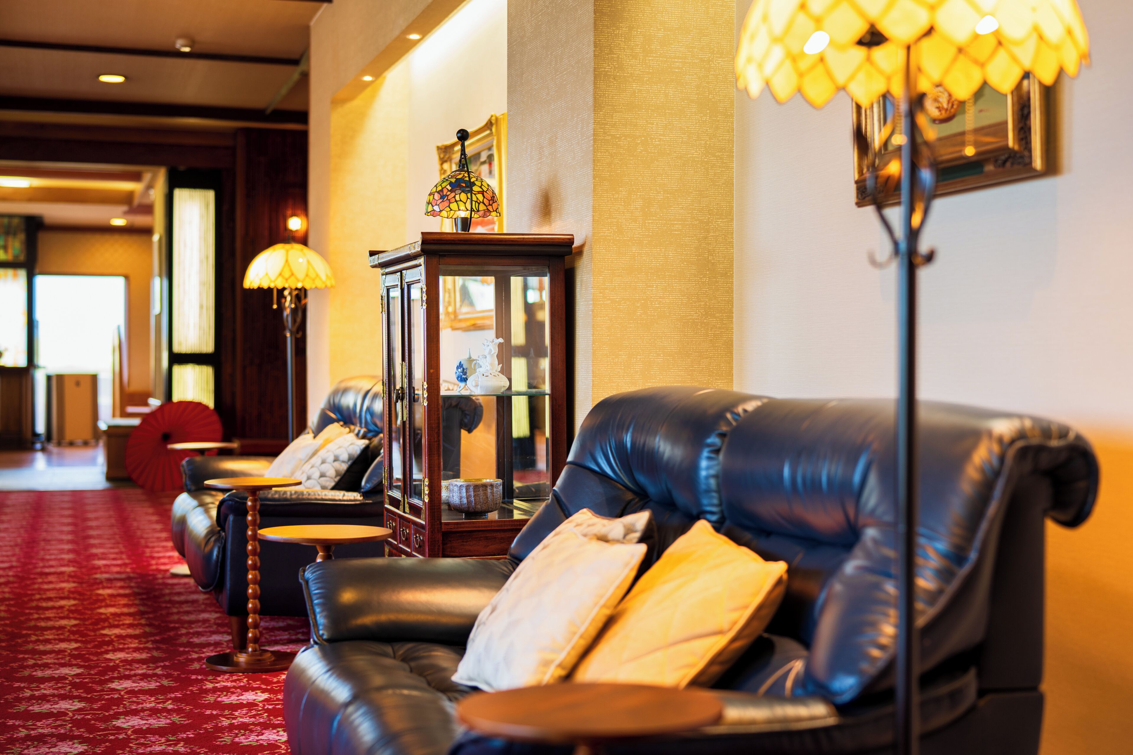 Um saguão de hotel com poltronas de couro, um tapete vermelho e um abajur com cúpula de vitral.