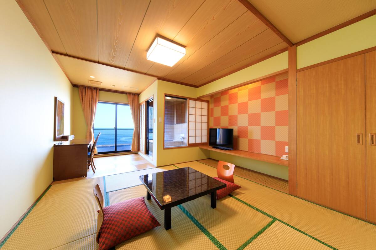 A traditional Japanese room with tatami flooring, a low table, a television, and sliding doors leading to a balcony.